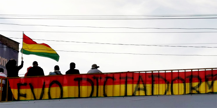 A protester in Bolivia waves a flag in front of graffiti saying 'Evo dictator!'