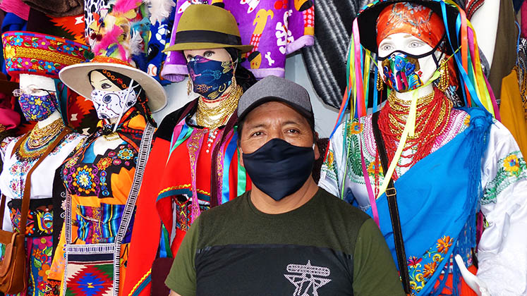 A market seller in a mask standing next to mannequins dressed in national costumes of Ecuador with protective masks in Cuenca