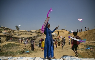 A woman dressed in blue holding up a kite in the centre foreground, taken on International Women's Day at a Rohingya refugee camp in Bangladesh