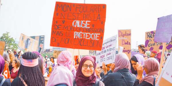 Hijabi holding protest sign reading "mon foulard n'est pas la cause de mon oppression mais le pretexte de mon exclusion"