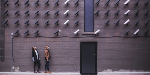 Two women on a street looking up at multiple rows of security cameras, all of which are pointed at them