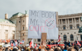 Man holding protest sign reading "My body my choice" with a crossed out syringe drawn