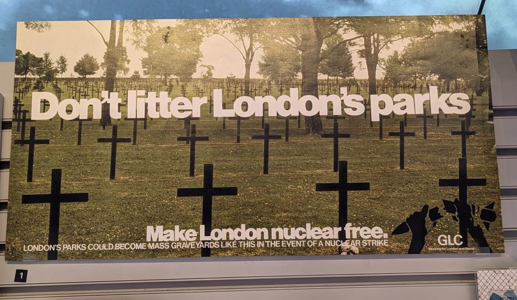 A photograph of a landscape poster that reads ‘Don’t litter London’s parks’. Beneath this is written ‘Make London nuclear free. London’s parks could become mass graveyards like this in the event of a nuclear strike’. Behind the text is a picture of crosses representing gravestones in a green field. 