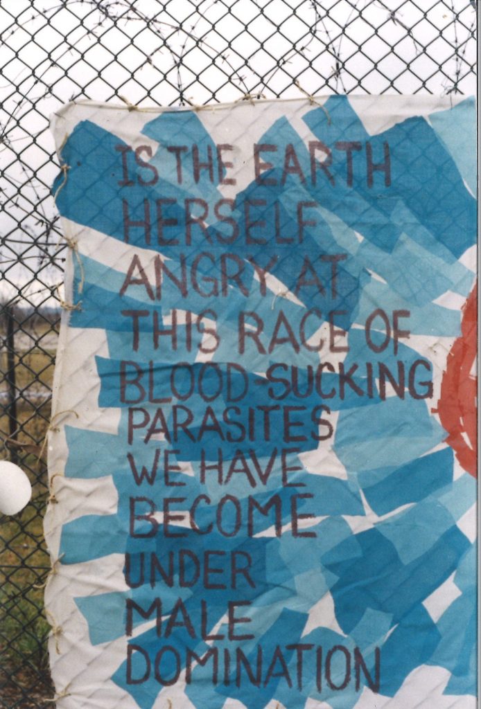 A photograph of a banner tied to a wire fence. The banner reads “Is the earth herself angry at this race of blood-sucking parasites we have become under male domination” in black letters against a blue and white background. 