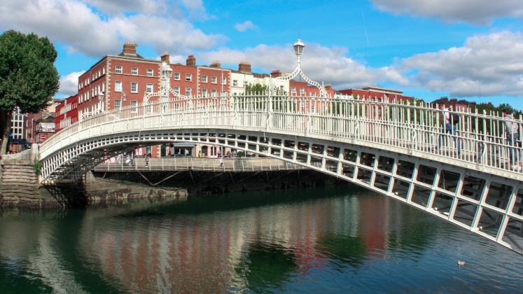 Image of the Ha'penny Bridge in Dublin Ireland which crosses the River Liffey, redbrick buildings and blue sky in the background.
