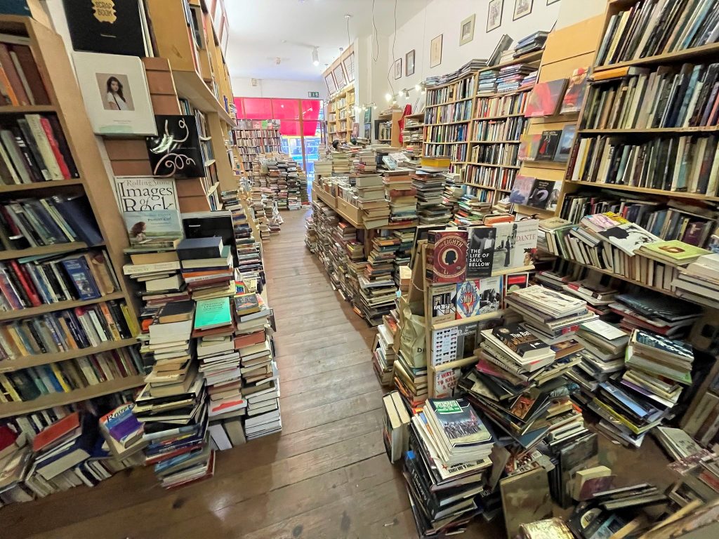 The interior of The Last Bookshop on Camden Street, Dublin. showing hap-hazard stacks of books on shelves and on a wooden floor.