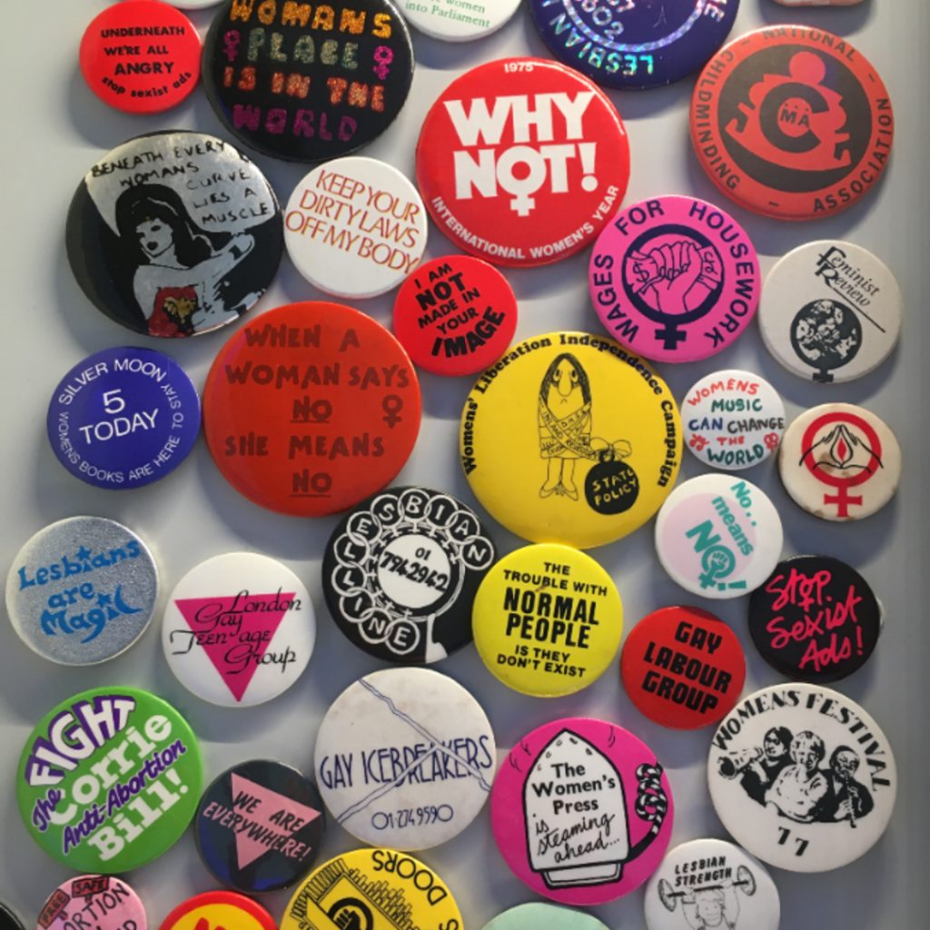 Colourful protest badges on a table all mixed up together and photographed from above. They include statements on like 'Lesbians are Magic' and 'Wages for Housework'