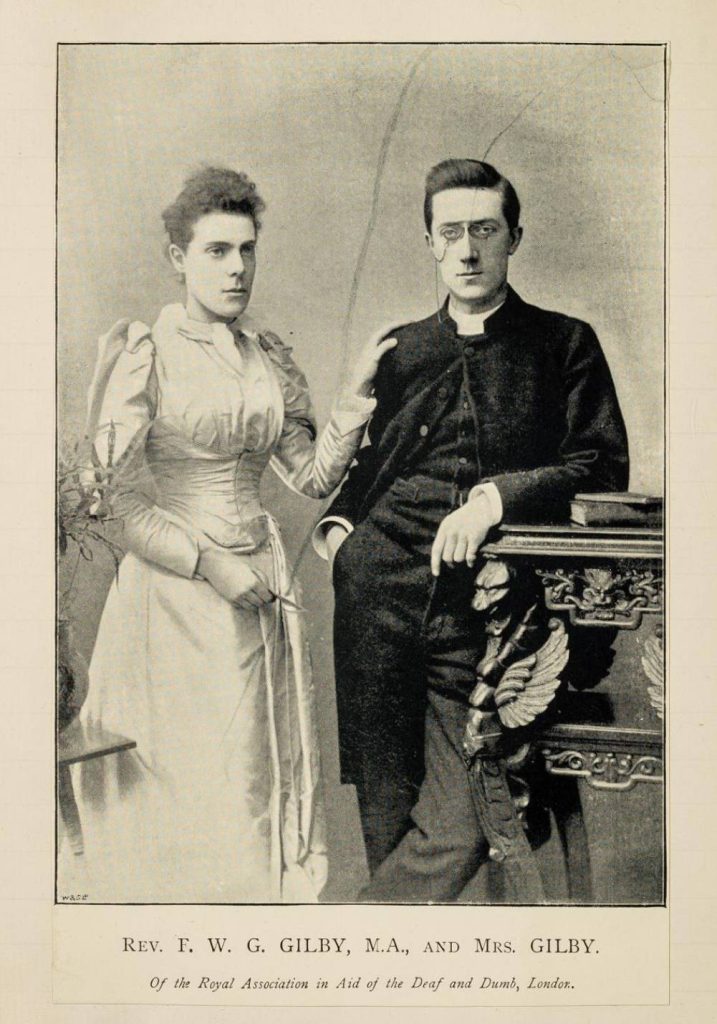 A photo of Gilby wearing spectacles and leaning on a shelf with a book on. Next to him is his wife wearing a white dress and with her hand on her husband's shoulder. 