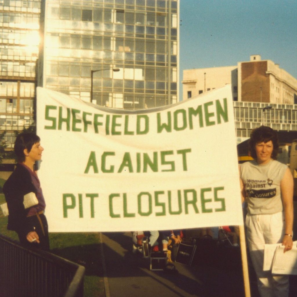 Two people stood near Sheffield bus station with a banner that reads "Sheffield Women Against Pit Closures". 