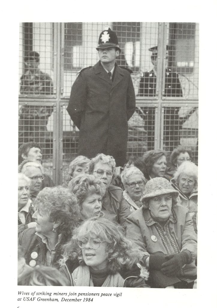 A photo in a book of wives of striking miners joining a pensioners' peace vigil at USAF Greenham, December 1984. Behind the seated women is a police officer stood in front of a metal gate. 