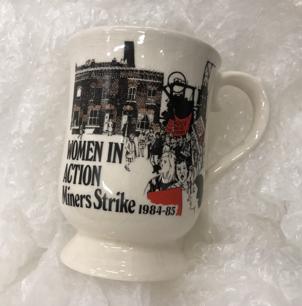 A mug with 'Women In Action Miners Strike 1984-85" written on it along with a drawing of a coal mine and people protesting on.