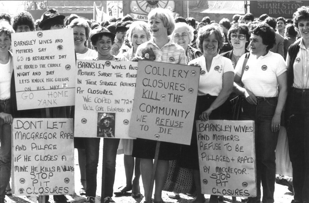 A group of mostly women smiling and holding placards. One reads as follows: "Colliery closures kill the community. We refuse to die". 