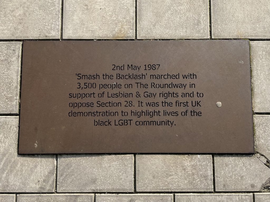 A plaque in the pavement. Reads as follows: 2nd May 1987 'Smash the Backlash' marched with 3,500 people on The Roundway in support of Lesbian & Gay rights and to oppose Section 28. It was the first UK demonstration to highlight lives of the black LGBT community. 